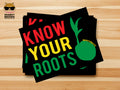 A stack of black stickers with the phrase 'KNOW YOUR ROOTS' in large red, yellow, and green letters, plus a green coconut sprout graphic, displayed on a light wood background. A Snarky Brown Studio logo appears in the top-left corner.